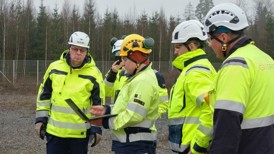 A group of men in high-visibility clothing stand together and look at a laptop.