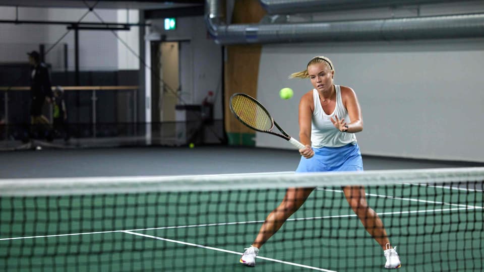 Kvinna som spelar tennis inomhus, hon står vid nät och ska till att slå en volley.
