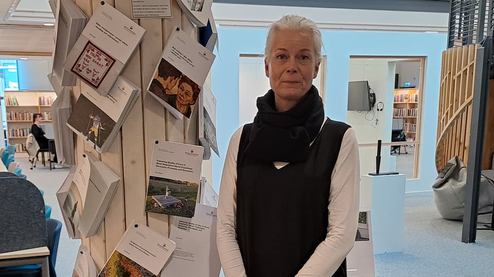 Ljushårig kvinna klädd i vit långärmad tröja med en svart väst över, runt halsen har hon en svart halsduk.
