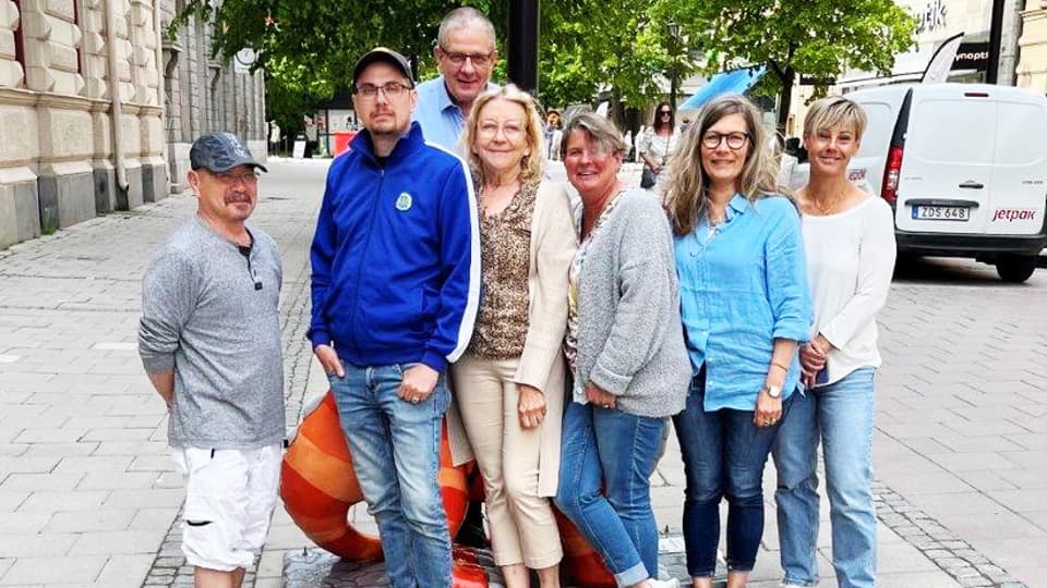 A group of people are standing together on a sidewalk, looking at the camera.