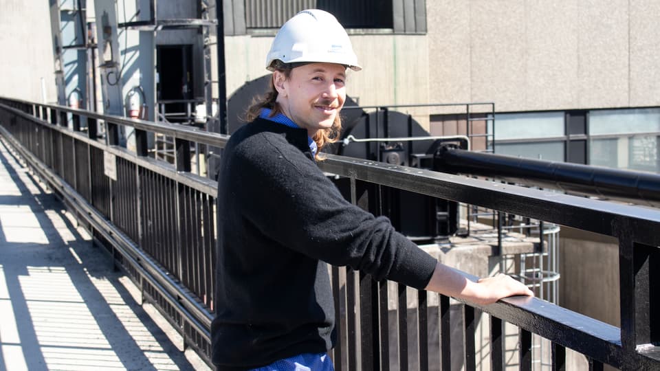 En man i en vit hjälm står vid ett svart staket i en industrimiljö.