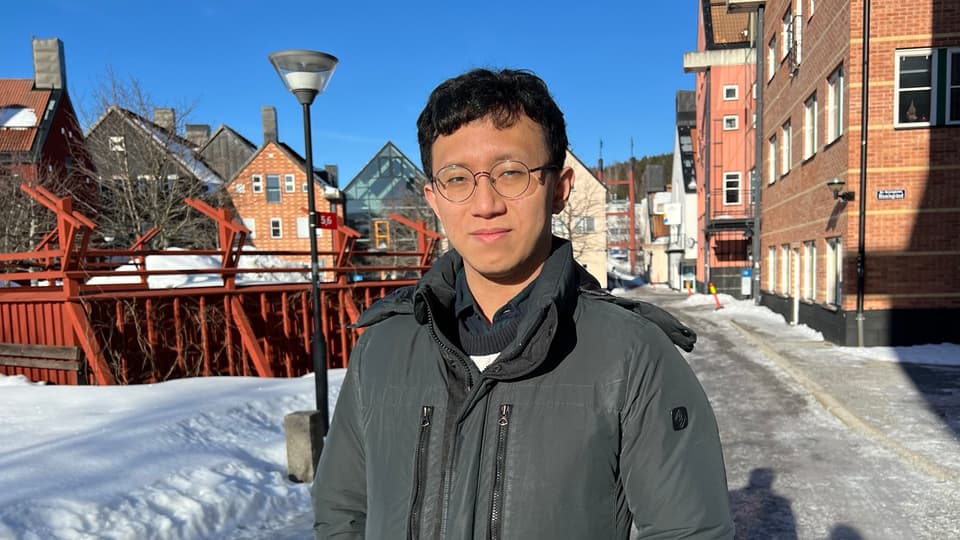 A man with dark hair and glasses stands outside in the sunshine. Campus buildings in snow and blue sky in the background.