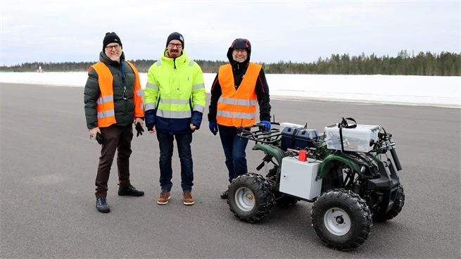 Three researchers in alert clothing stand by an autonomous quadricycle on a runway