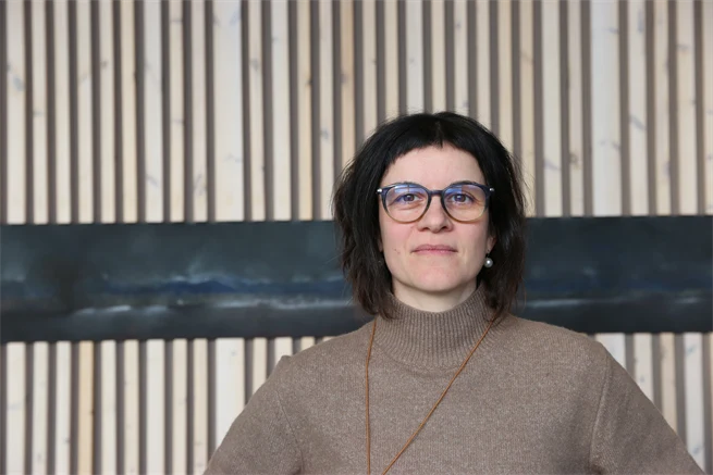 Serious woman with glasses in front of wooden panel.