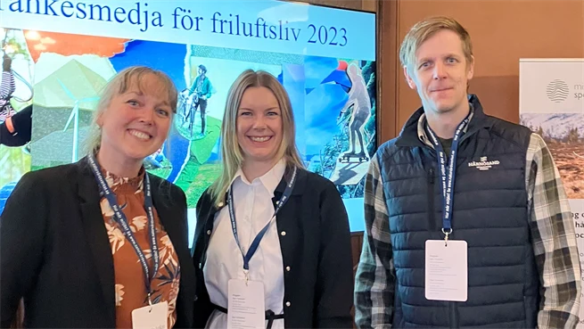Two women and a man stand in a room and look into the camera. Behind them is a screen with a presentation on it and they have name tags hanging around their necks
