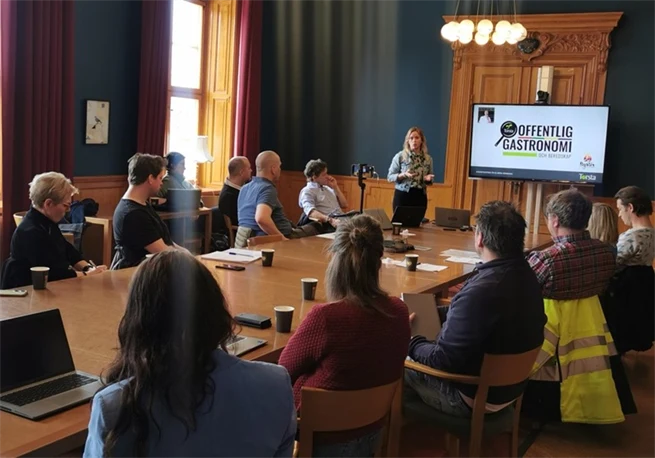 Two dozen people in a room with a woman at the front presenting in front of a screen