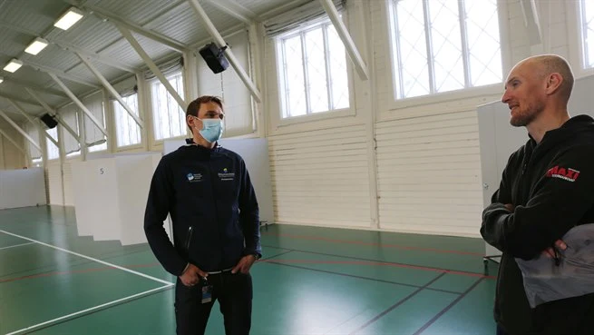 Två män står och pratar med varandra i en sporthall, en av männen bär munskydd.